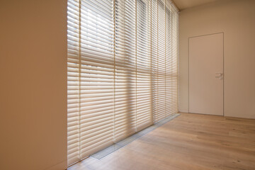 Motorized wood blinds in the interior. Automatic venetian blinds beige color on large windows. Wooden slats 50mm wide. A door to the room is near the window. 