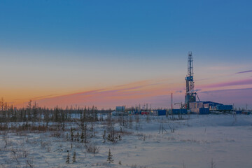 Winter landscape of a snowy forest tundra with a drilling rig for drilling an oil and gas well in the northern oil and gas field. Polar day sunset. Beautiful bright sky and clouds - obrazy, fototapety, plakaty