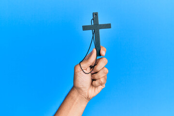 Hand of hispanic man holding christian cross over isolated blue background.