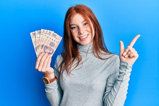 Young red head girl holding 500 mexican pesos banknotes smiling happy pointing with hand and finger to the side