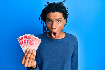 Young african american man holding 20 israel shekels banknotes scared and amazed with open mouth...