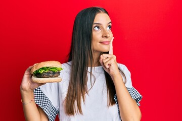 Young hispanic woman eating hamburger serious face thinking about question with hand on chin, thoughtful about confusing idea