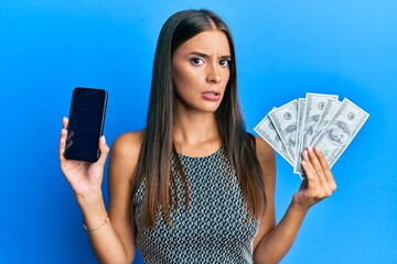 Young hispanic woman holding dollars and using smarpthone clueless and confused expression. doubt...