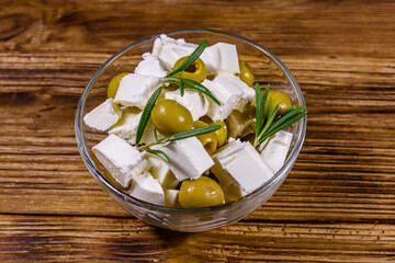 Chopped feta cheese, rosemary and olives in glass bowl on a wooden table