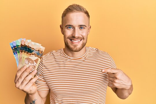 Young caucasian man holding canadian dollars smiling happy pointing with hand and finger
