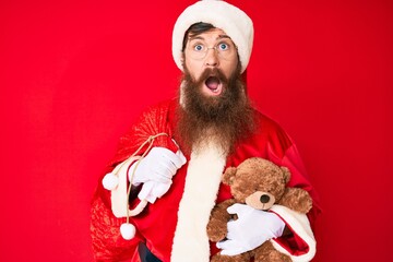 Handsome young red head man with long beard wearing santa claus costume holding teddy bear in shock face, looking skeptical and sarcastic, surprised with open mouth