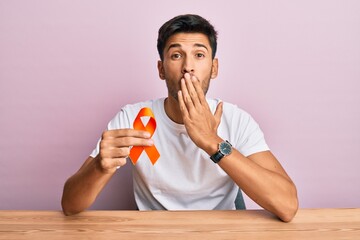 Young handsome man holding awareness orange ribbon covering mouth with hand, shocked and afraid for...
