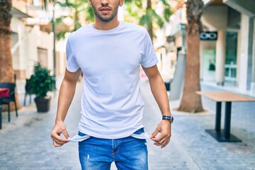 Young latin man with worried expression showing empty pockets walking at the city.