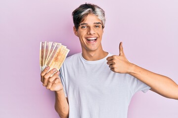 Young hispanic man holding 500 norwegian krone banknotes smiling happy and positive, thumb up doing...