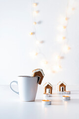 Cup of tea with gingerbread house and candles on white background. Christmas lights. Vertical frame