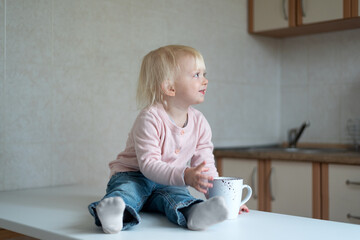 Blond child in the kitchen with cup in his hands. Portrait of toddler in the kitchen