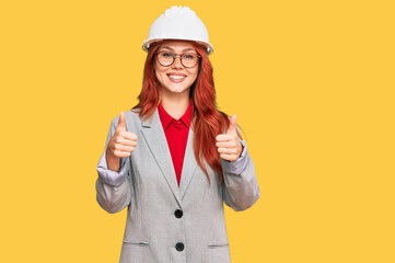 Young redhead woman wearing architect hardhat success sign doing positive gesture with hand, thumbs up smiling and happy. cheerful expression and winner gesture.