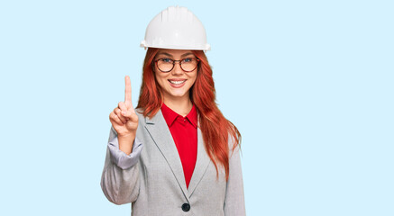 Young redhead woman wearing architect hardhat showing and pointing up with finger number one while smiling confident and happy.