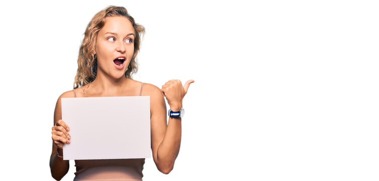 Beautiful caucasian woman holding blank empty banner pointing thumb up to the side smiling happy with open mouth