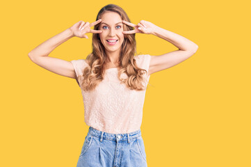 Young beautiful caucasian woman with blond hair wearing casual clothes doing peace symbol with fingers over face, smiling cheerful showing victory