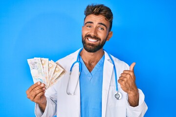 Young hispanic man wearing doctor uniform holding uk pounds banknotes smiling happy and positive, thumb up doing excellent and approval sign