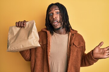 African american man with braids holding take away paper bag celebrating achievement with happy smile and winner expression with raised hand