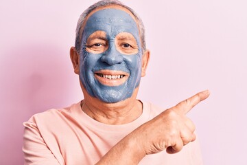 Senior man with grey hair wearing mud mask smiling cheerful pointing with hand and finger up to the...