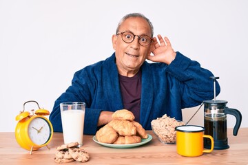 Senior handsome man with gray hair sitting on the table eating breakfast in the morning smiling with hand over ear listening and hearing to rumor or gossip. deafness concept.