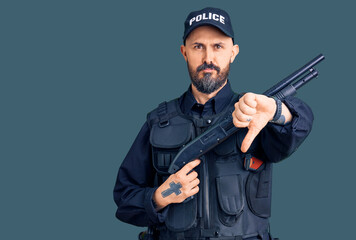 Young handsome man wearing police uniform holding shotgun with angry face, negative sign showing dislike with thumbs down, rejection concept