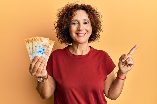 Beautiful middle age mature woman holding 10 swiss franc banknotes smiling happy pointing with hand and finger to the side