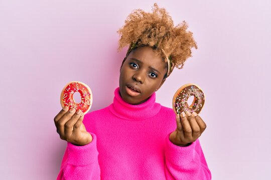 Young african woman with afro hair holding tasty colorful doughnuts in shock face, looking skeptical and sarcastic, surprised with open mouth