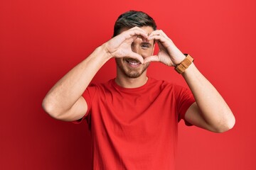 Handsome caucasian man wearing casual red tshirt doing heart shape with hand and fingers smiling looking through sign