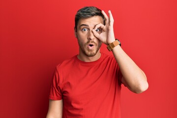 Handsome caucasian man wearing casual red tshirt doing ok gesture shocked with surprised face, eye looking through fingers. unbelieving expression.
