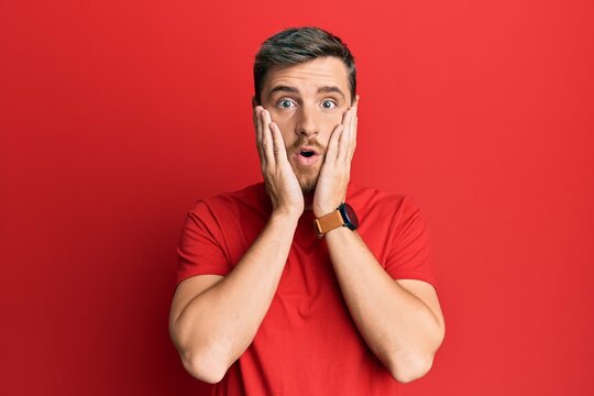 Handsome caucasian man wearing casual red tshirt afraid and shocked, surprise and amazed expression with hands on face