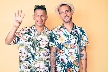 Young gay couple of two men wearing summer hat and hawaiian shirt showing and pointing up with fingers number four while smiling confident and happy.