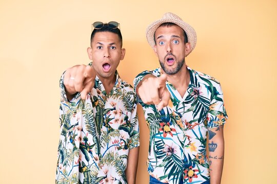Young gay couple of two men wearing summer hat and hawaiian shirt pointing with finger surprised ahead, open mouth amazed expression, something on the front