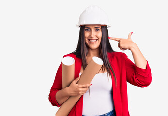 Young beautiful brunette woman wearing hardhat holding architect blueprint pointing finger to one self smiling happy and proud