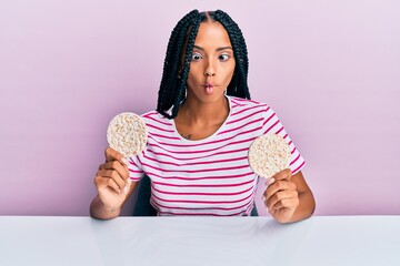 Beautiful hispanic woman eating healthy rice crackers making fish face with mouth and squinting eyes, crazy and comical.