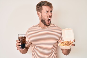 Young caucasian man eating fast food angry and mad screaming frustrated and furious, shouting with anger. rage and aggressive concept.