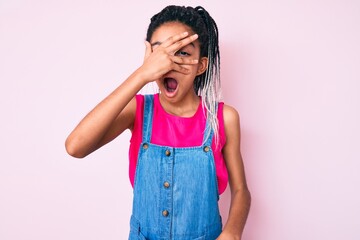 Young african american girl child with braids wearing casual clothes over pink background peeking in shock covering face and eyes with hand, looking through fingers with embarrassed expression.