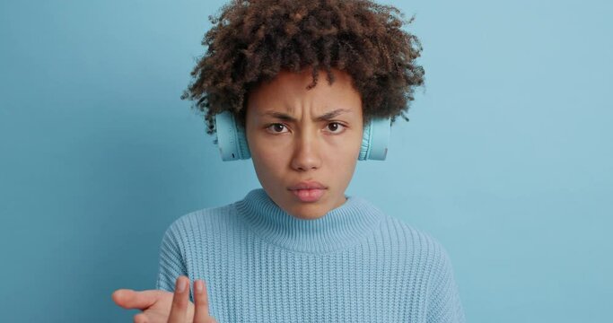 Perplexed dissatisfied dark skinned woman looks frustrated at camera says I dont agree has puzzled face expression dressed casually isolated over blue background listens music. Human emotions