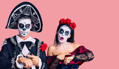 Young couple wearing mexican day of the dead costume over background in hurry pointing to watch time, impatience, upset and angry for deadline delay