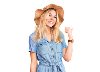 Young beautiful blonde woman wearing summer hat and dress smiling with happy face looking and pointing to the side with thumb up.