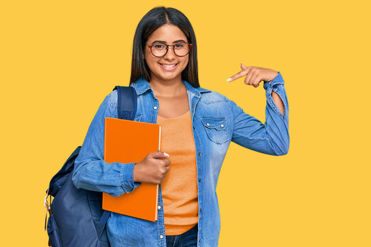 Young latin girl wearing student backpack and holding books looking confident with smile on face, pointing oneself with fingers proud and happy.
