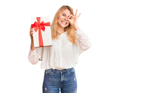 Young beautiful blonde woman holding gift doing ok sign with fingers, smiling friendly gesturing excellent symbol
