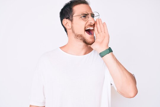 Young handsome man wearing casual clothes and glasses shouting and screaming loud to side with hand on mouth. communication concept.