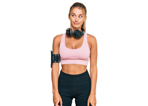 Young blonde woman wearing gym clothes and using headphones smiling looking to the side and staring away thinking.