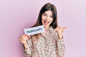 Young beautiful caucasian girl holding february word paper pointing thumb up to the side smiling happy with open mouth