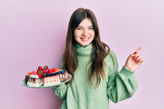 Young beautiful caucasian girl holding cake slices smiling happy pointing with hand and finger to the side