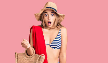 Young beautiful blonde woman wearing bikini and hat holding summer wicker handbag scared and amazed with open mouth for surprise, disbelief face