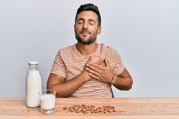 Handsome hispanic man drinking healthy almond milk smiling with hands on chest, eyes closed with grateful gesture on face. health concept.