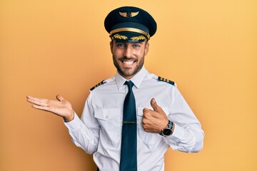 Handsome hispanic man wearing airplane pilot uniform showing palm hand and doing ok gesture with...