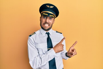 Handsome hispanic man wearing airplane pilot uniform pointing aside worried and nervous with both...