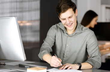 Fototapeta na wymiar Young businessman and programmer in a green shirt is writing some notes in his business plan, while he is sitting at the desk in a cabinet together with his female colleague on the background