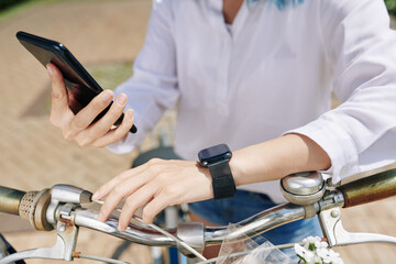 Close-up image of woman with smartwatch sitting on bicycle and checking applications on smartphone and smartwatch